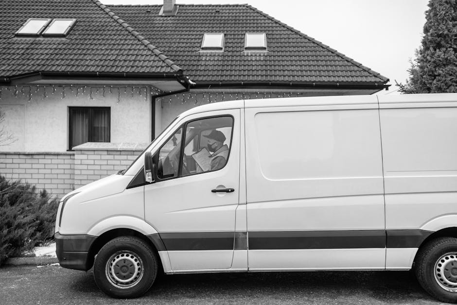 A white delivery van parked outside a residential property during daytime, with a man inside the van reading a document, possibly related to a house removal or home relocation. The van is positioned o
