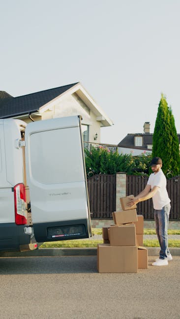 A white delivery van parked outside a residential property during daytime, with a man inside the van reading a document, possibly related to a house removal or home relocation. The van is positioned o