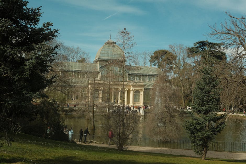 A large glass and stone Victorian-style greenhouse with a domed roof and classical columns, situated in Crystal Palace Park, visible through a park landscape with leafless trees and green grass in the