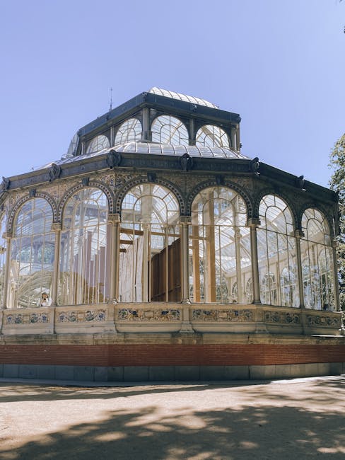 An ornate Victorian-style greenhouse constructed with intricate metal framework and large glass panels, situated outdoors under a clear blue sky. The structure features multiple arched windows and a c
